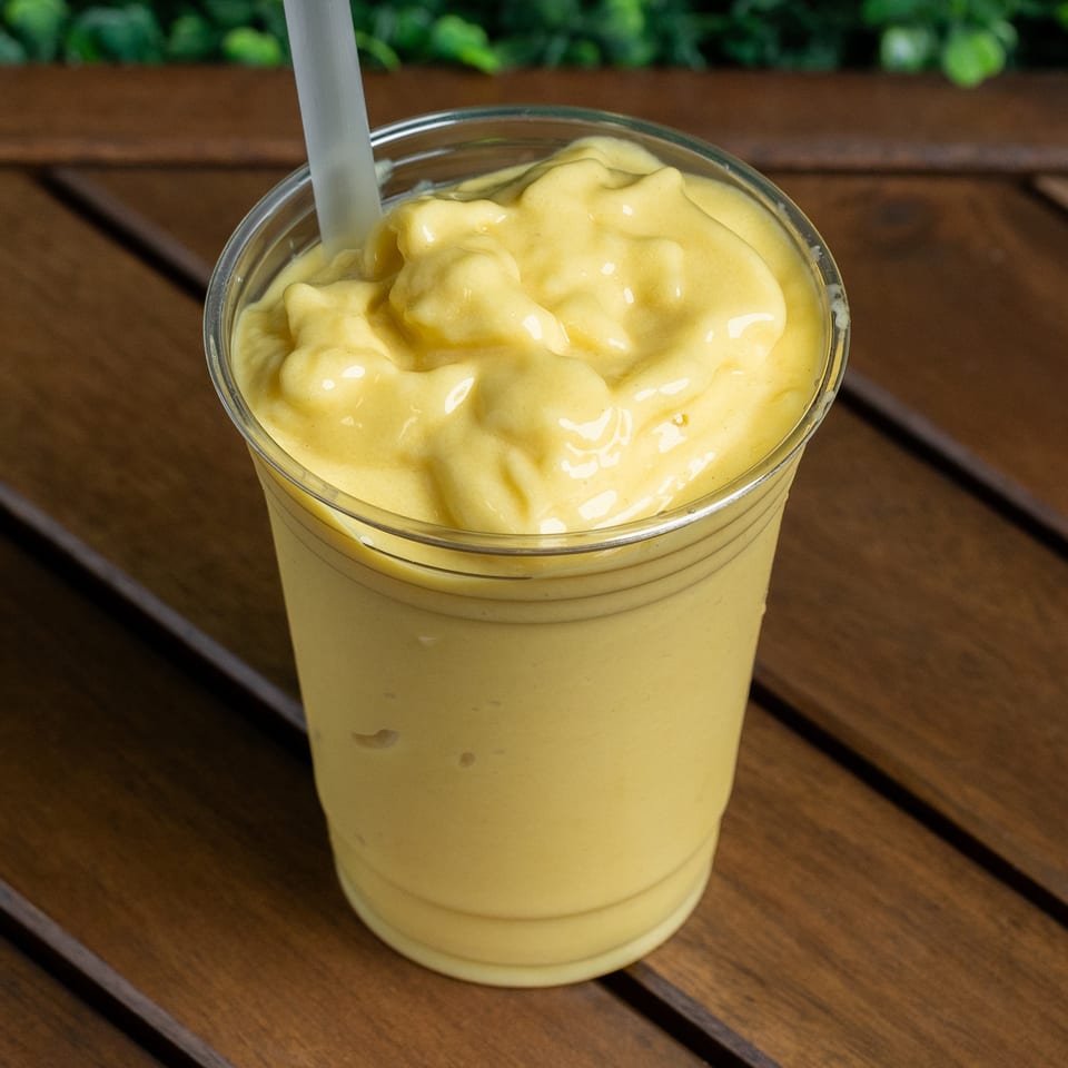 Creamy yellow mango smoothie in a clear plastic cup with a straw on a wooden table.