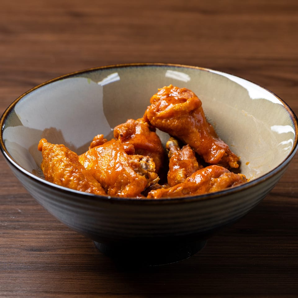 A bowl of saucy, orange-glazed chicken wings sits on a dark wooden table.