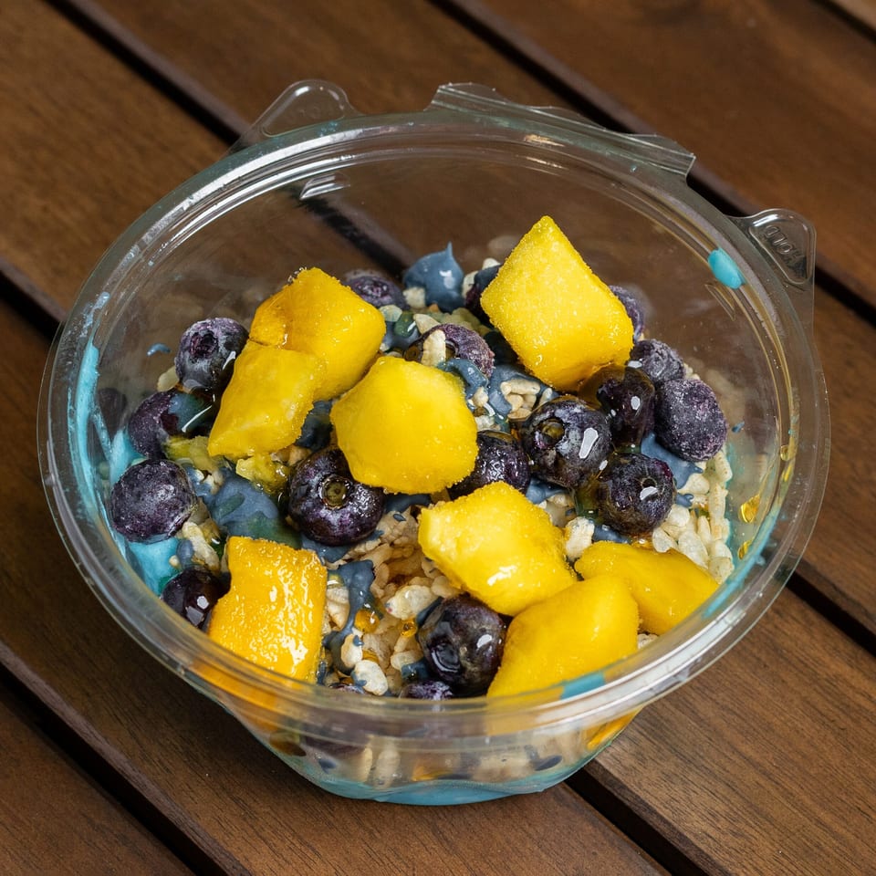 A clear plastic bowl filled with blue yogurt, blueberries, mango chunks, and rice cereal on a wooden table.