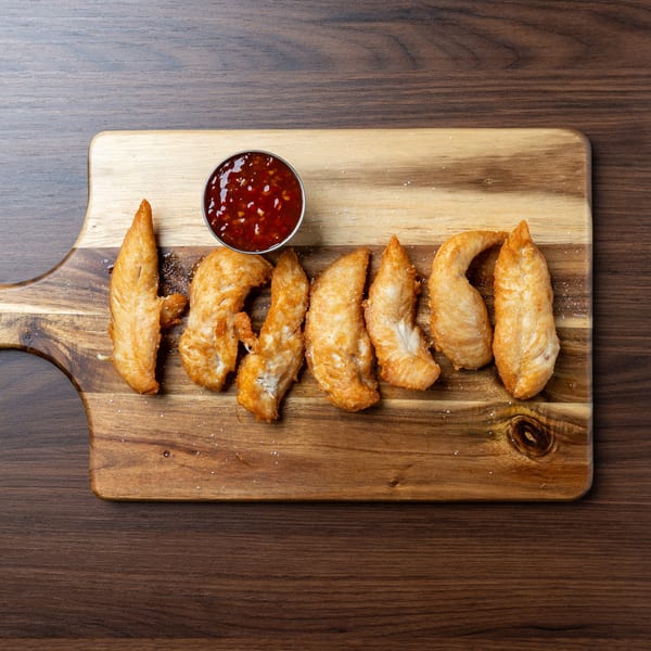 Golden-brown chicken tenders with a side of sweet chili dipping sauce on a wooden cutting board.
