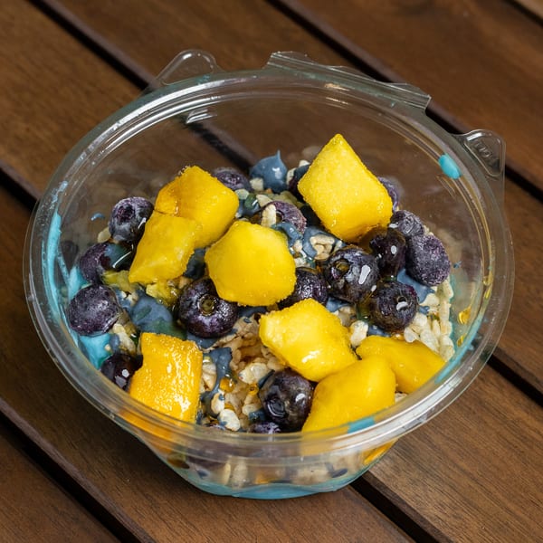 A clear plastic bowl of blue yogurt, blueberries, mango chunks, and rice cereal on a wooden table.